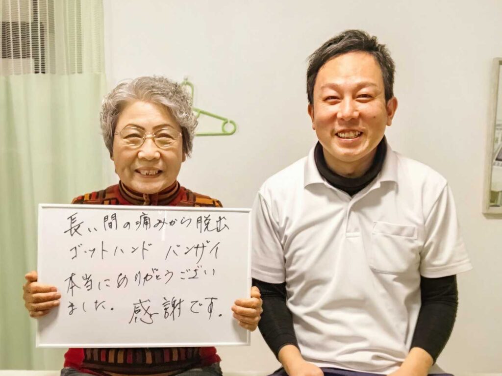 「長い間の痛みから脱出、ゴットハンド バンザイ 本当にありがとうございました。感謝です。」と書かれたホワイトボードを持つ都城市在住の70代女性の患者様と、笑顔の「はる整体院」院長の写真