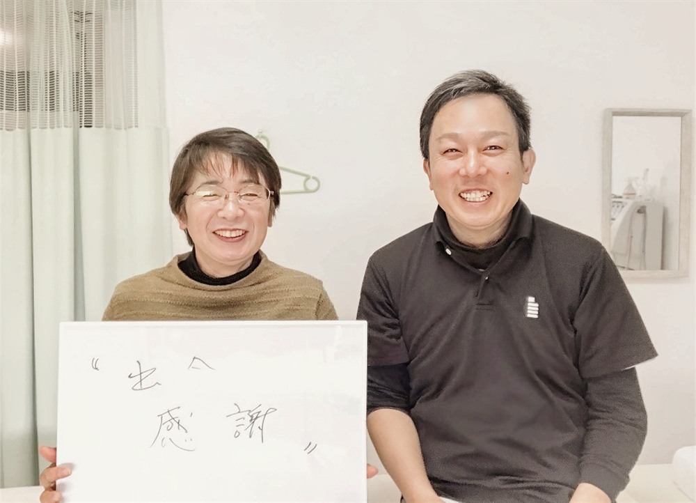 「出会い 感謝」と書かれたホワイトボードを持つ都城市在住の60代女性患者様と、笑顔の院長の写真