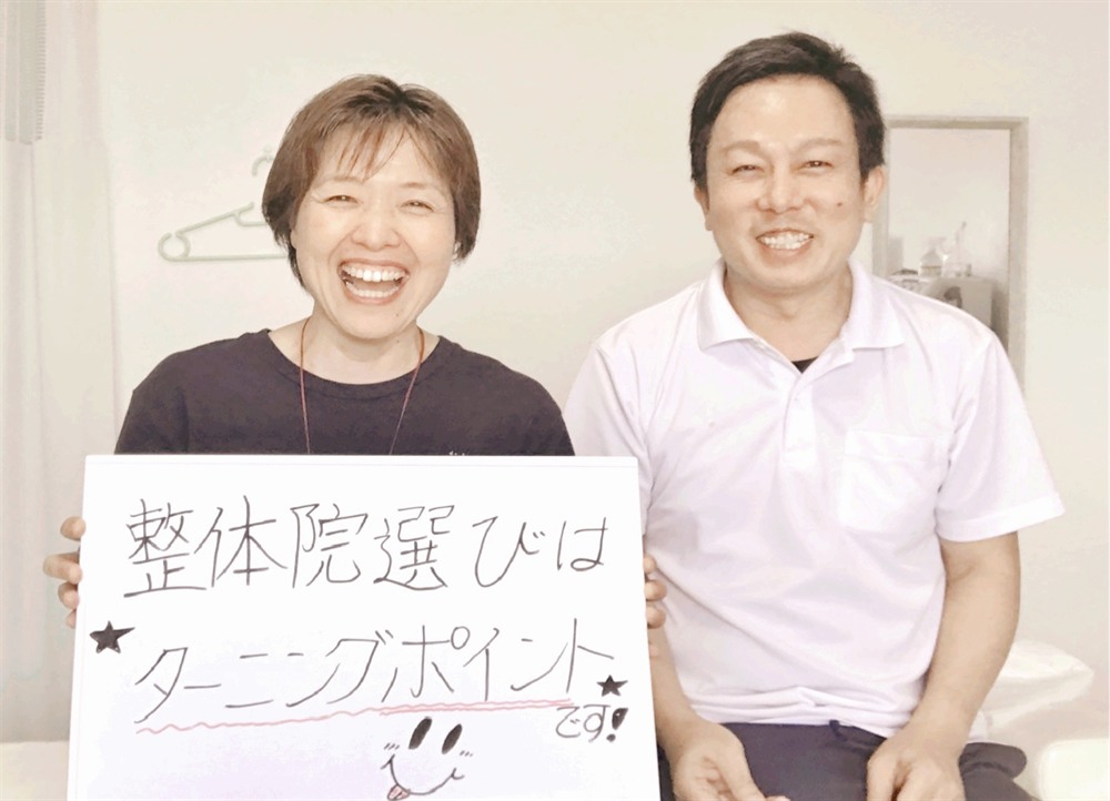 「整体院選びはターニングポイントです！」と書かれたホワイトボードを持つ都城市在住の50代の女性患者様と、笑顔の院長との写真